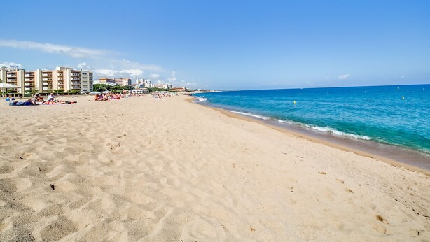 Una playa arenosa con un mar azul cristalino, edificios al fondo y algunas personas relajándose.