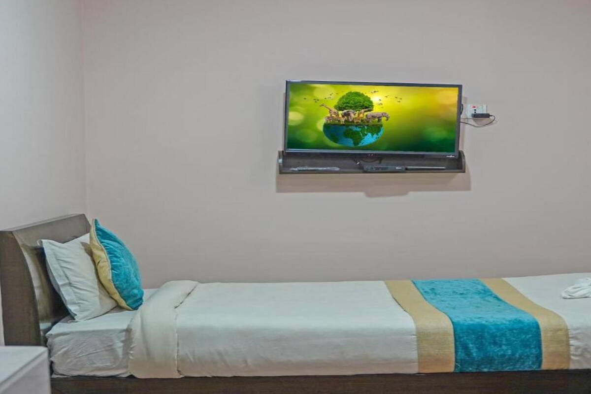 Una cama individual con colcha azul y blanca, un televisor montado en la pared y una mesita de noche con lámpara.