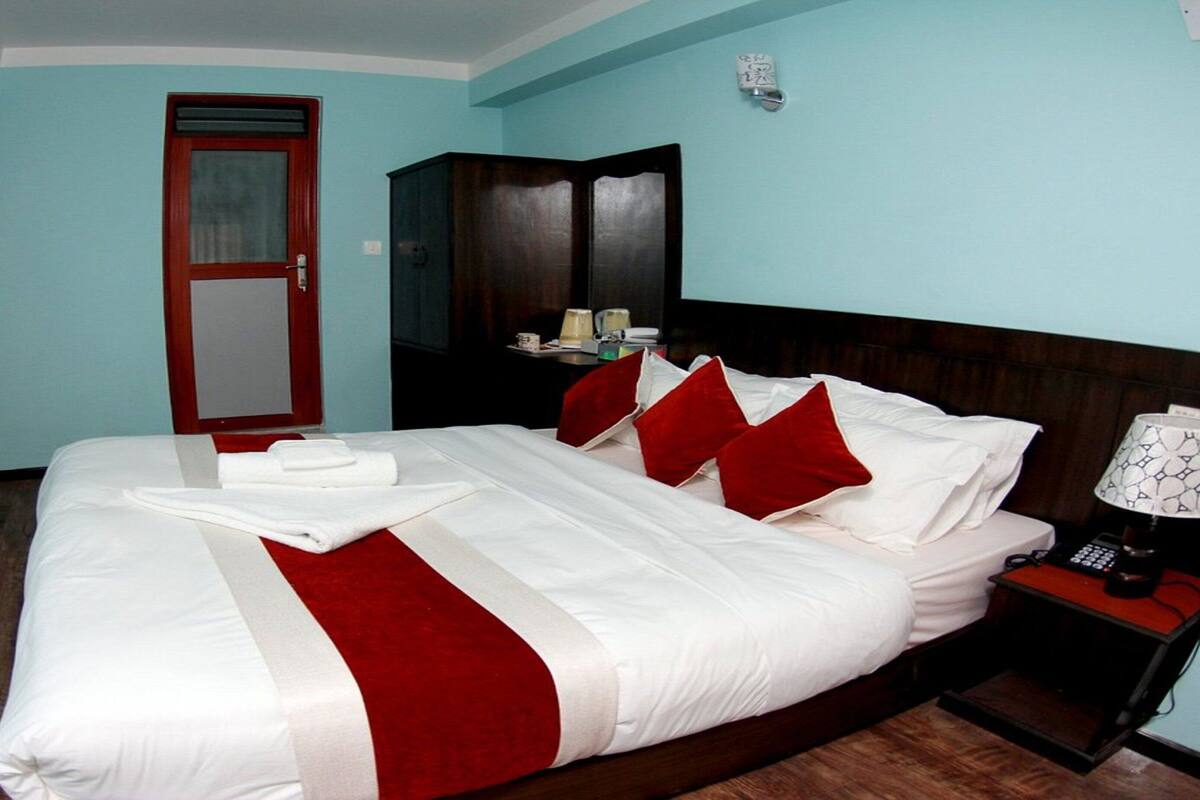 Una cama bien hecha con ropa de cama blanca y roja, un cabecero de madera, una mesita de noche con una lámpara y un armario en una habitación con paredes azul claro.