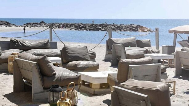 Outdoor seating with cushions and tables on a sandy beach, with the ocean and rocky outcrops in the background. - Grande Villa Avec Piscine Proche mer + Bateau (Mireval)