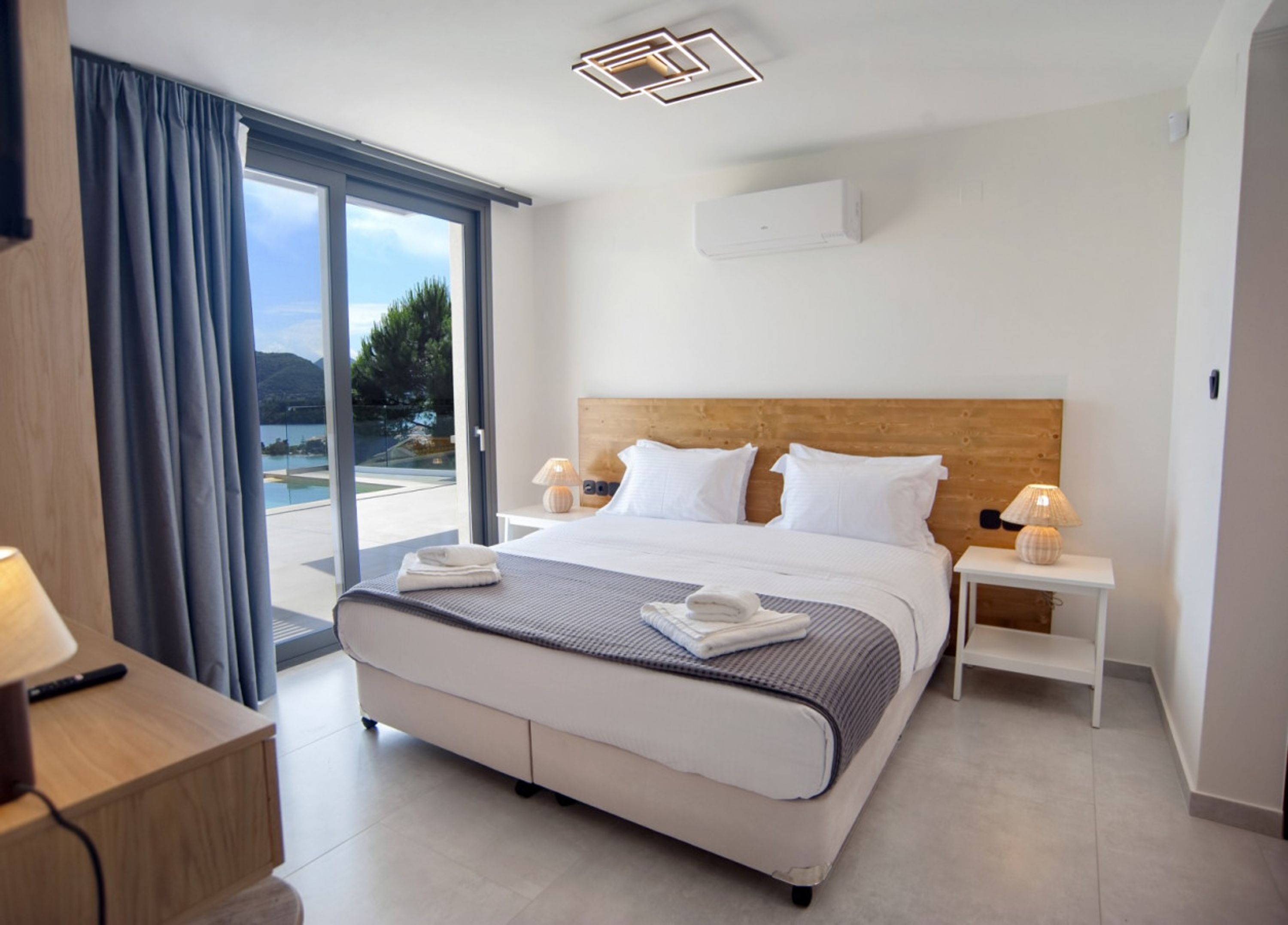 Una camera d'albergo moderna con un letto grande, una testiera in legno, due comodini con lampade e vista sull'esterno tramite una porta scorrevole in vetro.