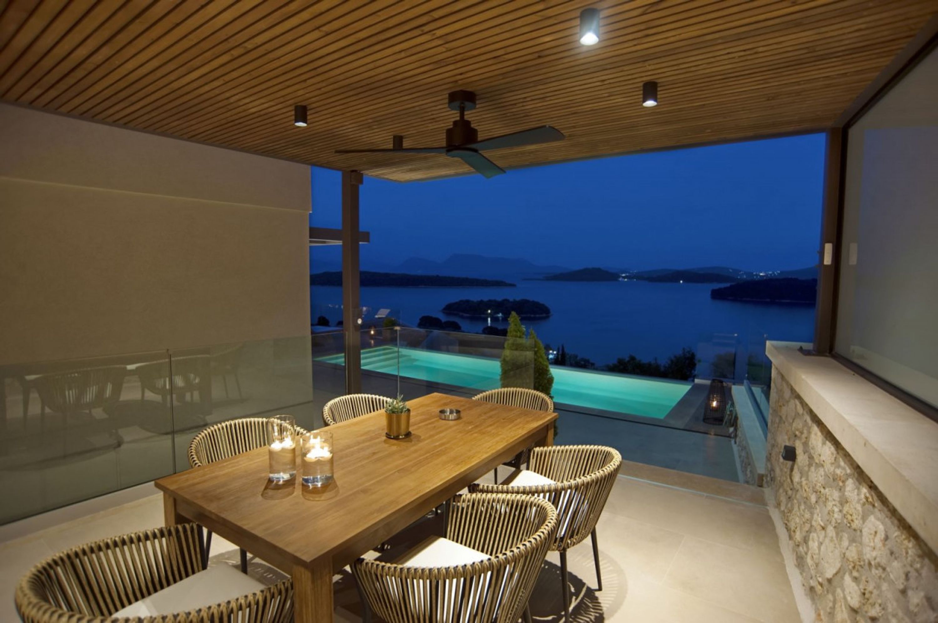 Un espace repas avec une table en bois et des chaises en osier, donnant sur une piscine et un plan d’eau au crépuscule.