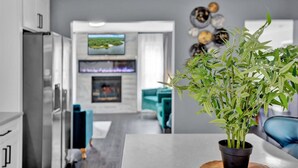 A modern kitchen with white cabinets, a dining area with a blue chair, and a living room with a TV and decorative wall art. - 6BR | Fire Pit, Games, Theater Room + EV Charger (Long Pond)