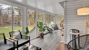 A covered porch with a view of a yard, featuring a dining table with chairs, a hanging chair, and a lamp. - 6BR | Fire Pit, Games, Theater Room + EV Charger (Long Pond)