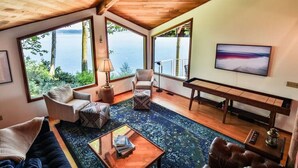 A spacious living room with large windows overlooking a body of water, a comfortable seating area, and a flat-screen TV mounted on the wall. - Family Friendly Water View Retreat w/ Sauna (Port Ludlow)