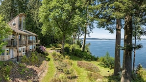 A wooden house with a deck overlooking a lake, surrounded by dense trees and greenery. - Family Friendly Water View Retreat w/ Sauna (Port Ludlow)