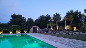 Una piscina con solárium de piedra, rodeada de jardines ajardinados y árboles.