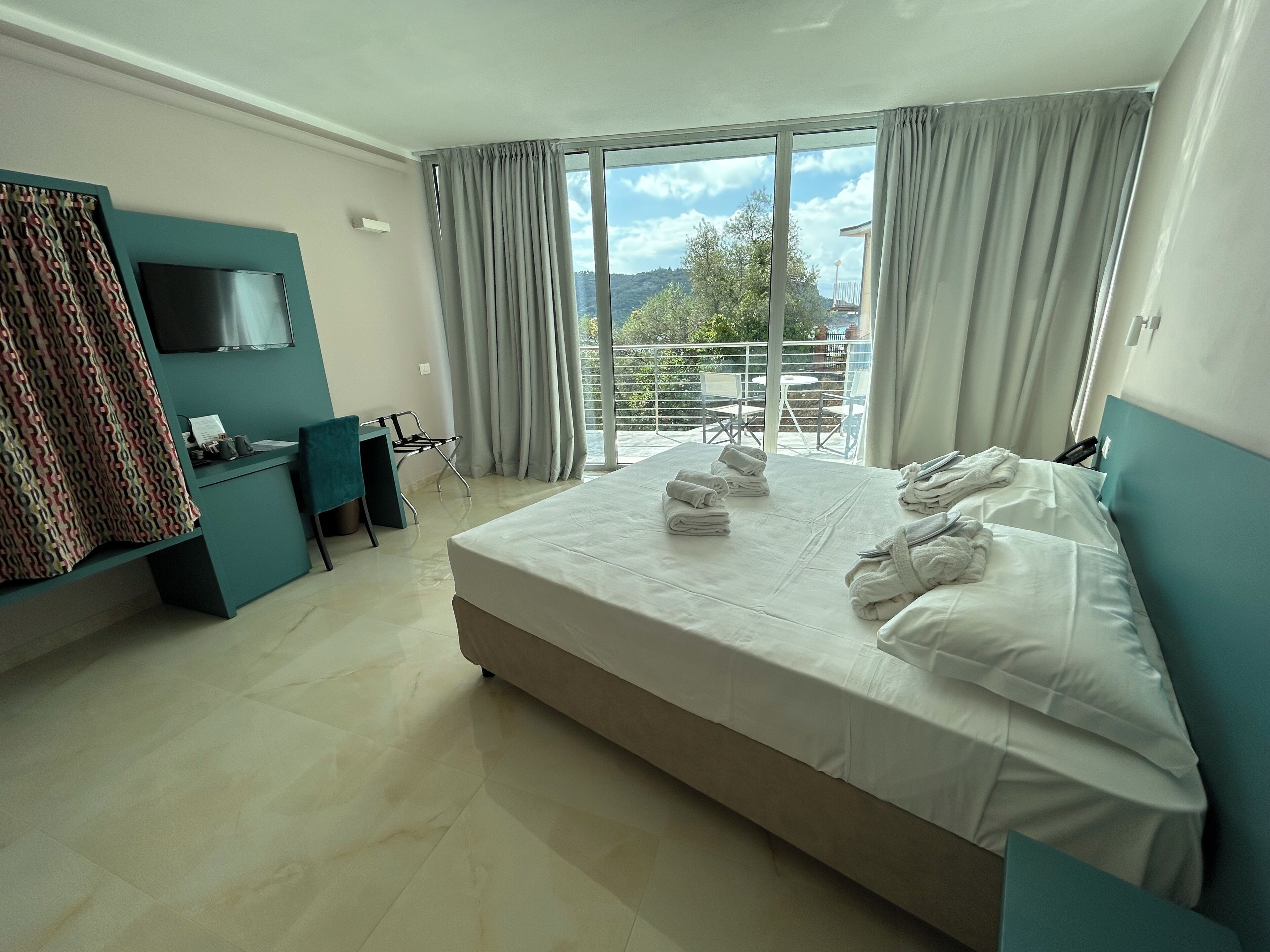 A hotel room with a large bed, a desk, a TV, and a balcony with a view of the outdoors.