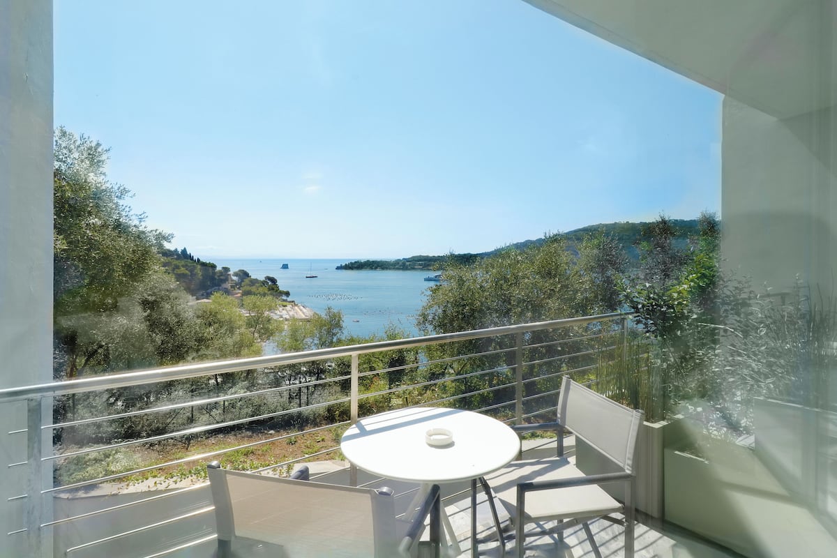 A balcony with a white table and chairs, overlooking a coastal landscape with trees and boats.