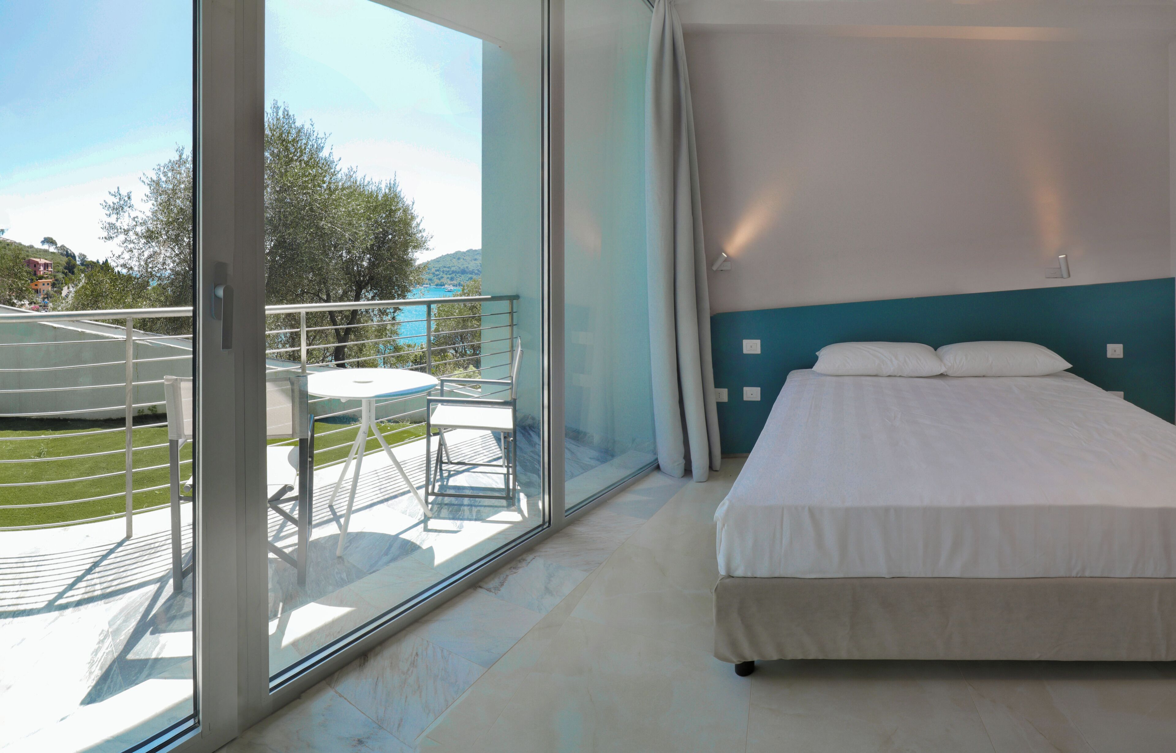 A bedroom with a large sliding glass door leading to a balcony, a bed with white bedding, and a view of a tennis court and trees.