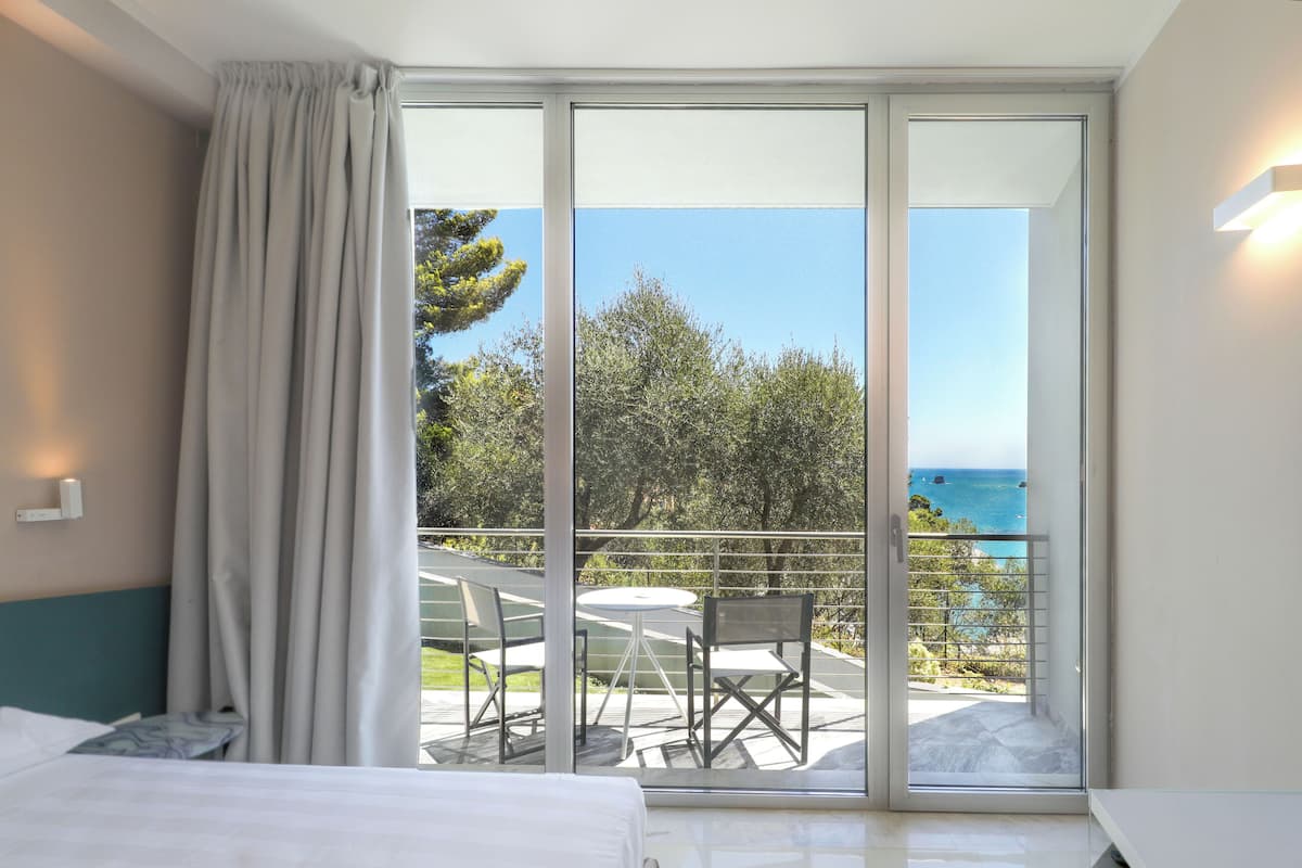 A hotel room with a large sliding glass door leading to a balcony with a table and chairs, overlooking a scenic view of the sea and greenery.