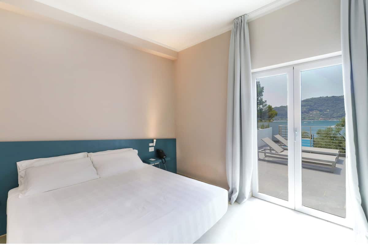 A hotel room with a large bed, a nightstand, a phone, and a view of a balcony with outdoor furniture.