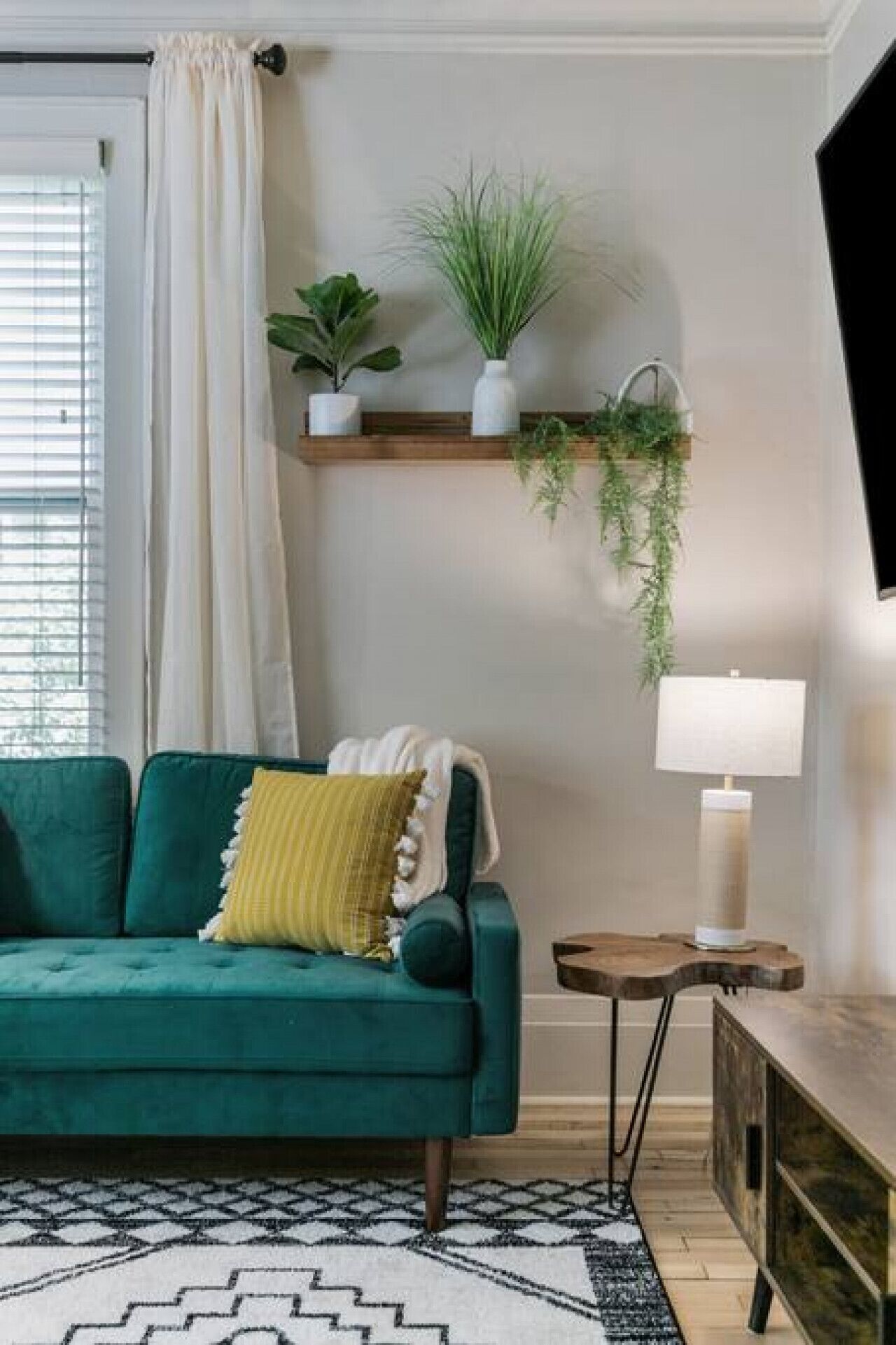 Ein blaugrünes Sofa mit einem gelben Kissen, ein kleiner Holztisch mit einer Lampe und ein Wandregal mit Pflanzen.