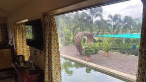 Una habitación con un ventanal grande, televisor, una mesa de madera con tetera y tazas, y vistas a un jardín con fuente y plantas.