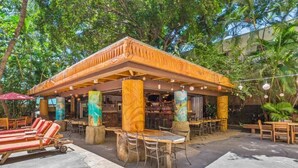 An outdoor restaurant with wooden tables and chairs, surrounded by palm trees and a covered seating area.