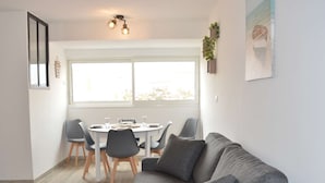 A modern living room with a grey sofa, a dining table with chairs, and a large window providing natural light. - Seaside Apartment: 2 Rooms, Sleeps 5, Modern, 50m from Beach, A/C, Parking Included (Narbonne)