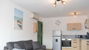 A compact living space with a grey sofa, a dining table, a kitchenette with a fridge, oven, and coffee machine, and a green door. - Seaside Apartment: 2 Rooms, Sleeps 5, Modern, 50m from Beach, A/C, Parking Included (Narbonne)