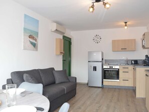 Um espaço de estar compacto com um sofá cinzento, uma mesa de jantar, uma cozinha compacta com frigorífico, forno e máquina de café, e uma porta verde.
