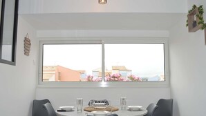 A dining area with a round table, four chairs, and a window with a view of buildings and pink flowers. - Seaside Apartment: 2 Rooms, Sleeps 5, Modern, 50m from Beach, A/C, Parking Included (Narbonne)