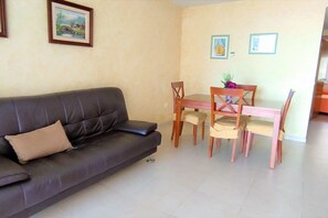 Una sala de estar con un sofá de color oscuro, una mesa de comedor de madera con sillas y dos cuadros en la pared.