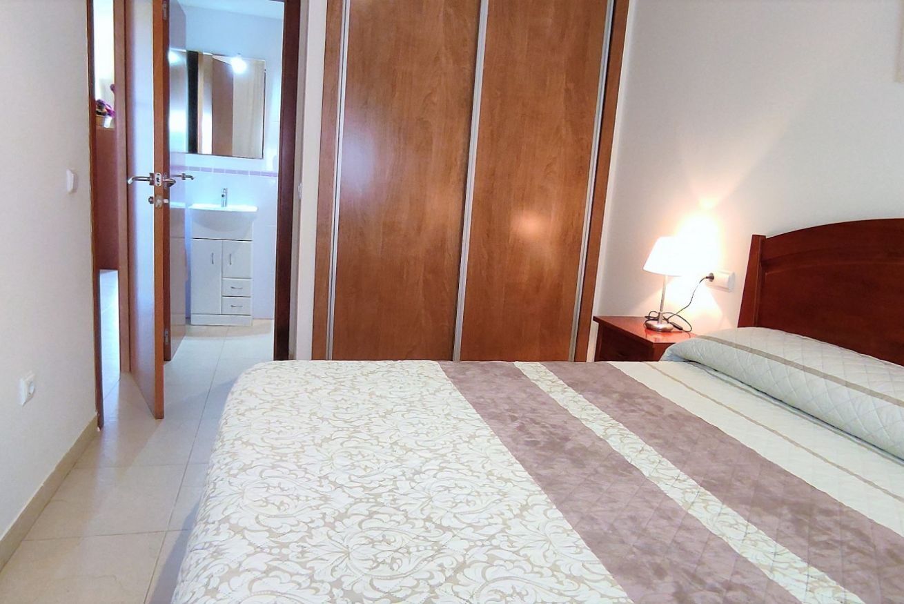 Une chambre à coucher avec un lit, une table de chevet avec une lampe, une armoire en bois et une salle de bain visible à travers une porte ouverte.