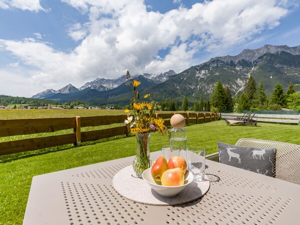 Une table sur laquelle se trouvent un saladier de fruits, un vase de fleurs et un verre d’eau, le tout placé devant un paysage de montagnes et une clôture en bois.