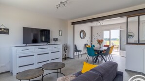 A modern living room with a grey sofa, yellow cushions, a flat-screen TV, and a dining area with blue chairs and a white table.