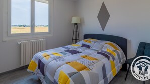 A bedroom with a large window, a bed with a geometric-patterned duvet, a bedside lamp, and a chair.