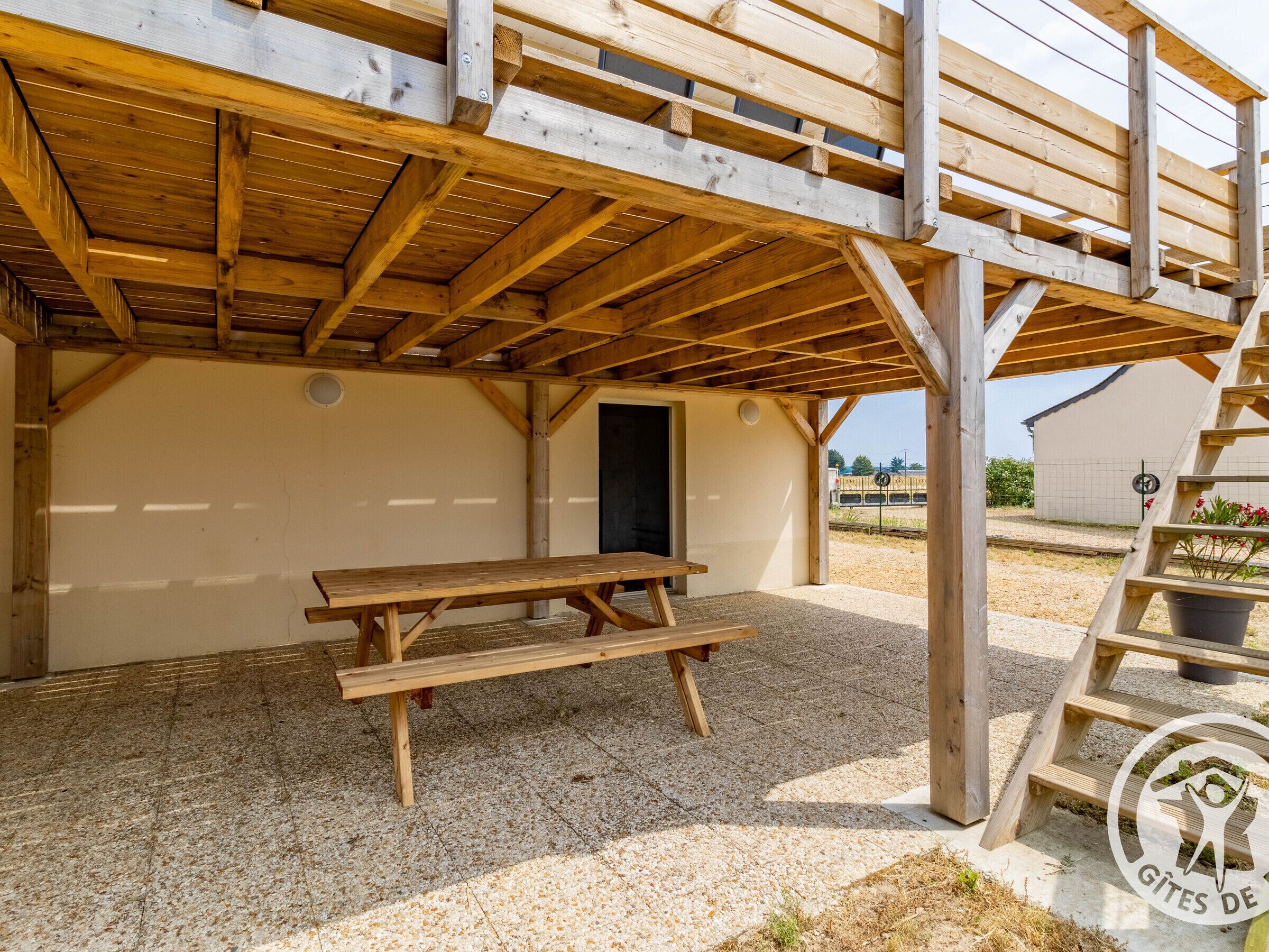 A wooden outdoor seating area with a picnic table, a covered section with a door, and a staircase leading to an upper level.
