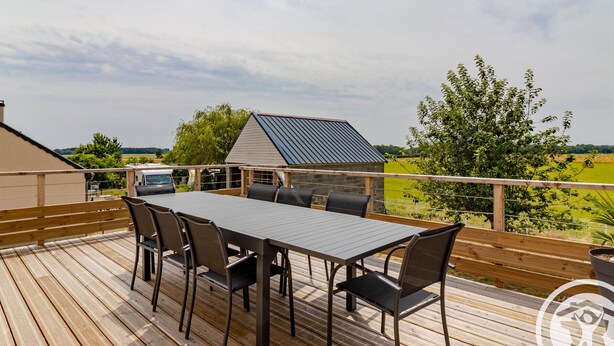A wooden deck with a table and chairs, overlooking a green landscape and a building with a solar panel.