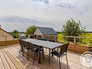 Une terrasse en bois avec une table et des chaises, offrant une vue sur un paysage verdoyant et un bâtiment équipé d’un panneau solaire.