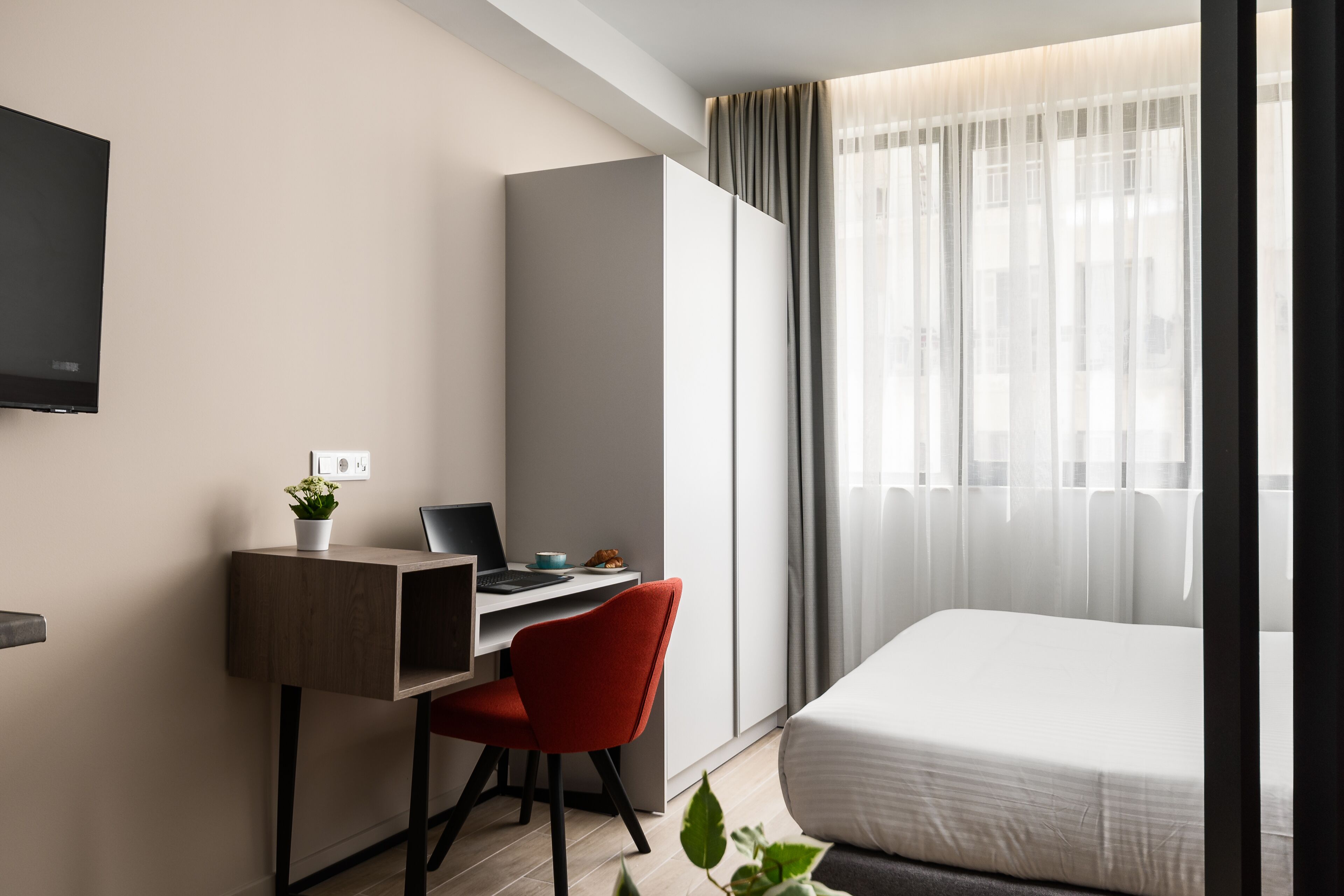 A modern hotel room with a bed, a desk with a laptop, a red chair, a TV mounted on the wall, and a window with sheer curtains.