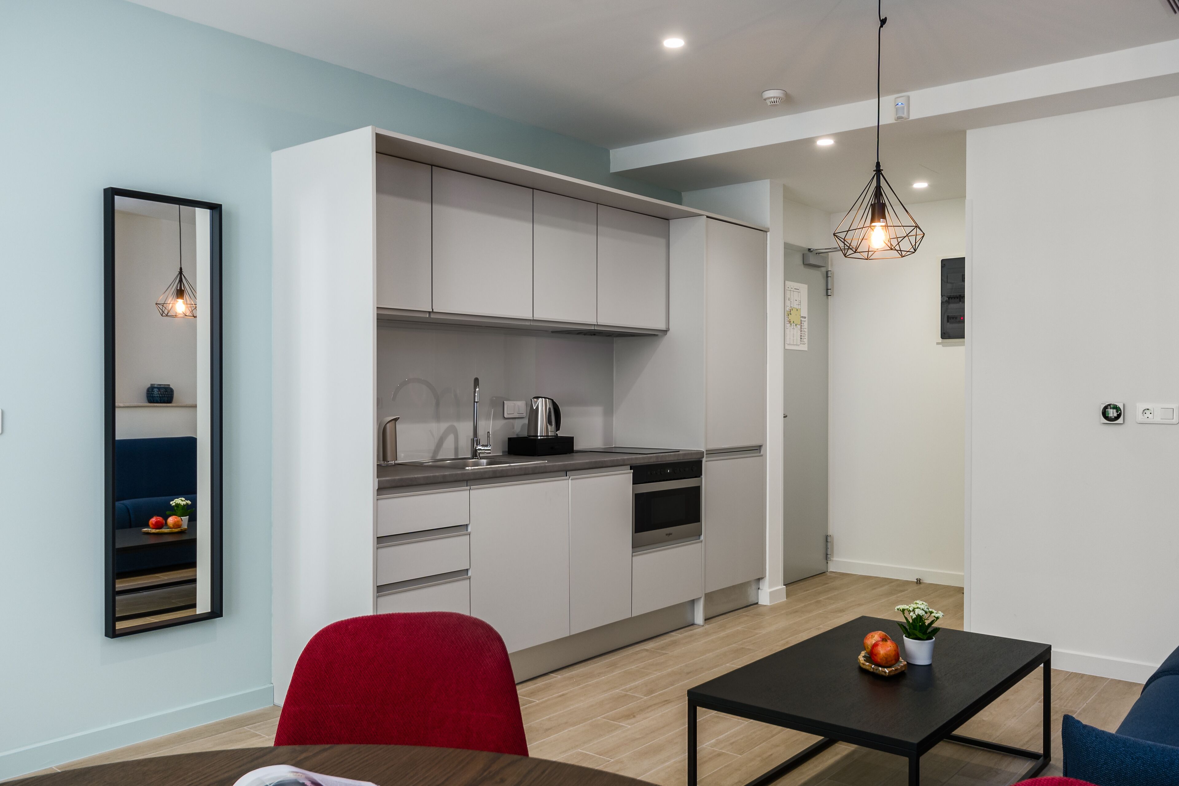 A modern kitchen with white cabinets, a black countertop, and a built-in oven. There is a red chair and a black table with a fruit bowl and a plant.