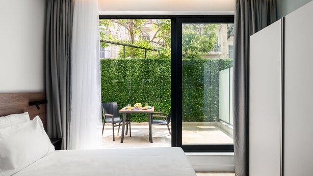 Ein modernes Hotelzimmer mit einem großen Bett, einem Essbereich mit Tisch und Stühlen sowie einer Glastür, die auf einen Balkon mit Blick auf Grünflächen und Gebäude führt.