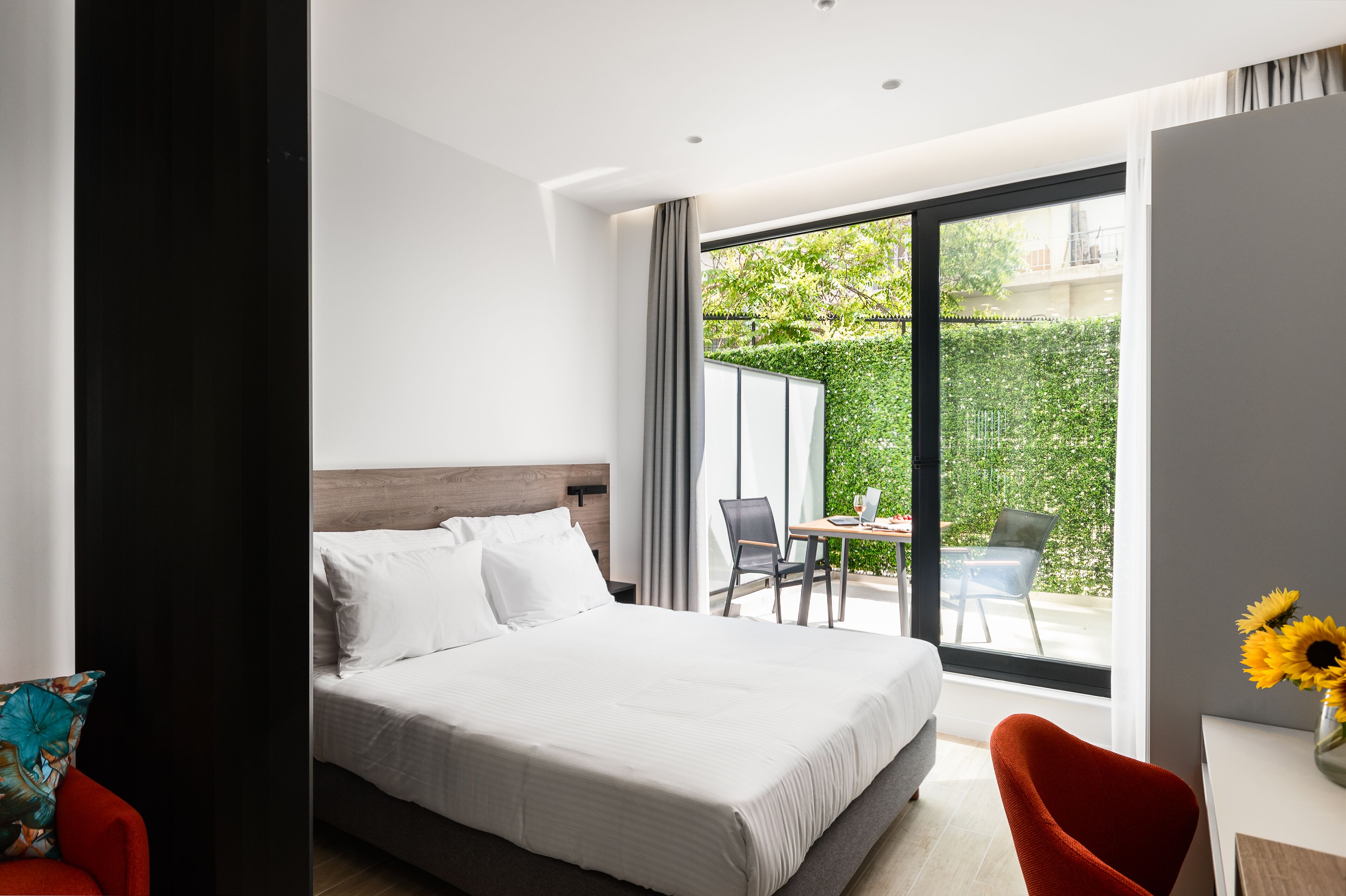 Una habitación de hotel moderna con una cama grande, un escritorio con silla y vistas a un jardín verde a través de una puerta corrediza de vidrio.