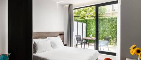 A modern hotel room with a large bed, a desk with a chair, and a view of a green garden through a sliding glass door.