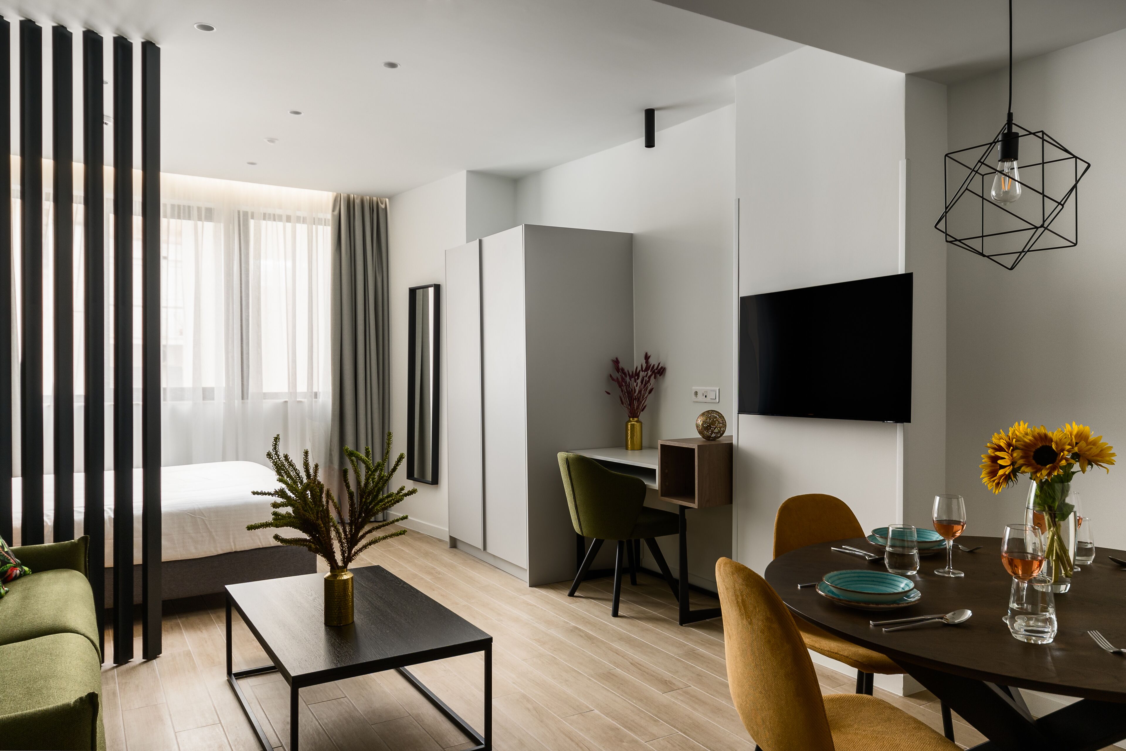 A modern living room with a dining area, a flat-screen TV mounted on the wall, and a large window with sheer curtains.