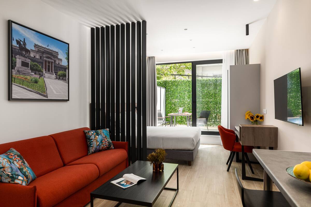 Een moderne woonkamer met een rode bank, een salontafel en een televisie. Er is een balkon met uitzicht op groen.