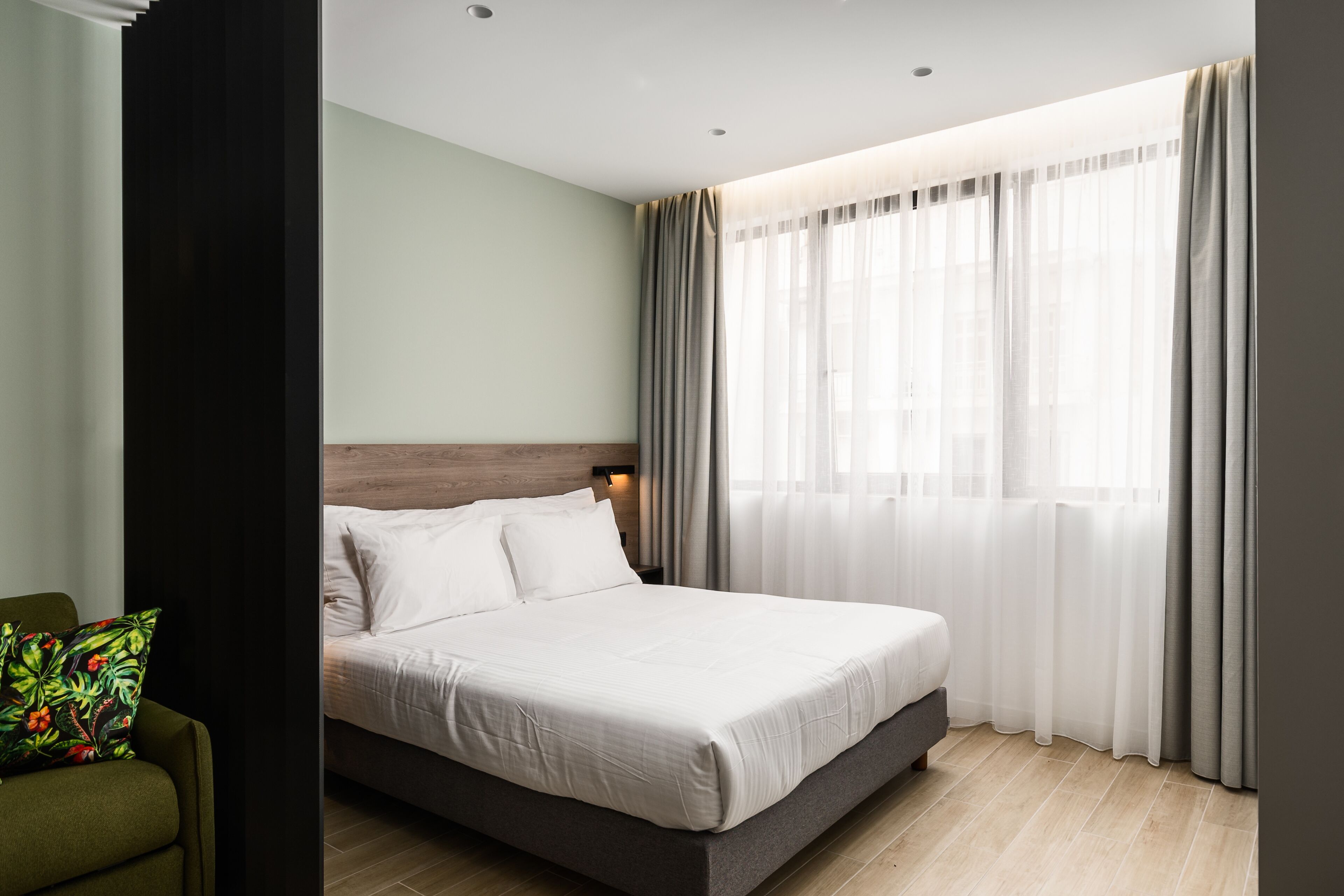 Quarto de hotel moderno com uma cama grande, uma poltrona verde e uma janela com cortinas leves.