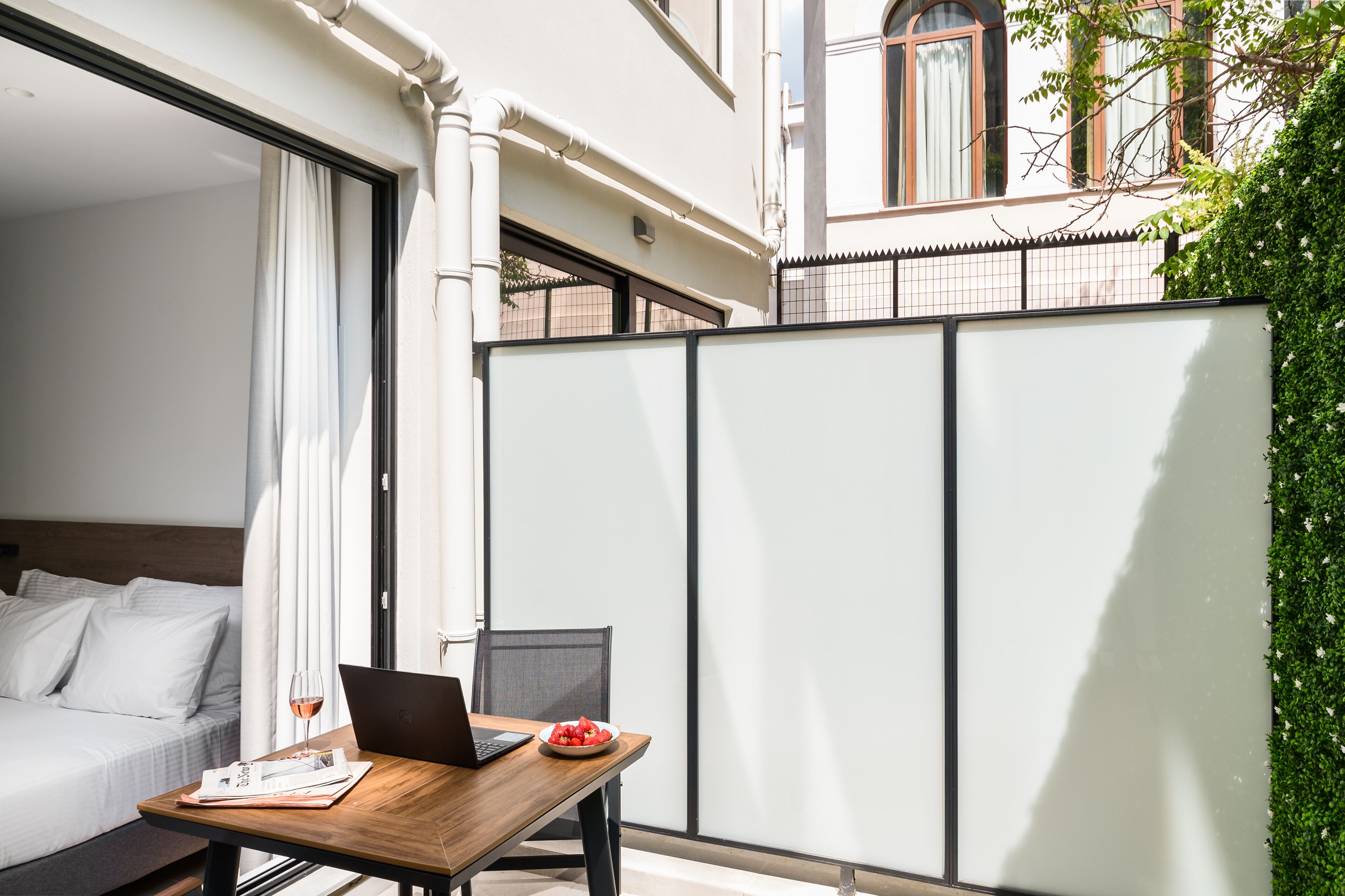 A hotel room with a bed, a wooden table with a laptop and a bowl of fruit, and a view of the outside.