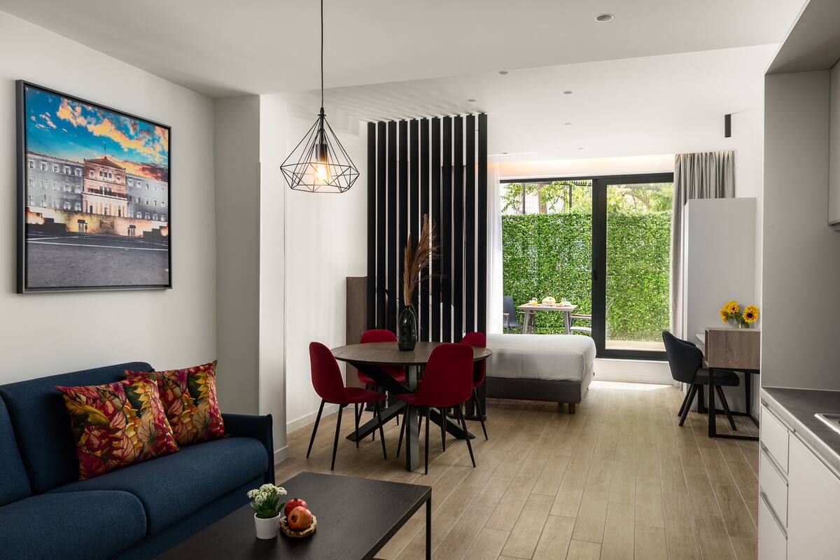 Een moderne woonkamer met een blauwe bank, rode stoelen, een eettafel en een groot raam met uitzicht op groen.