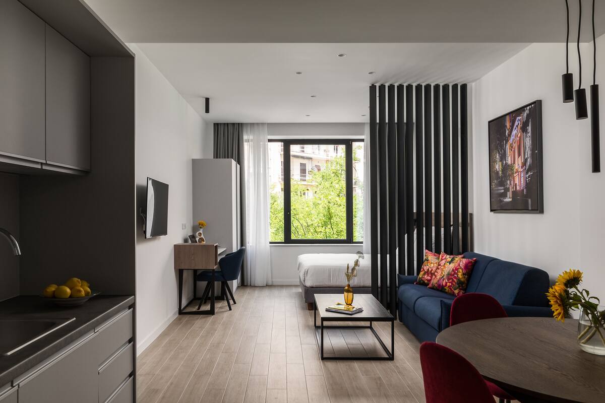 Een moderne woonkamer met een bank, een salontafel en een eettafel. Er is een keuken met keukenkastjes en een spoelbak. Een raam biedt uitzicht op het buitengebied.