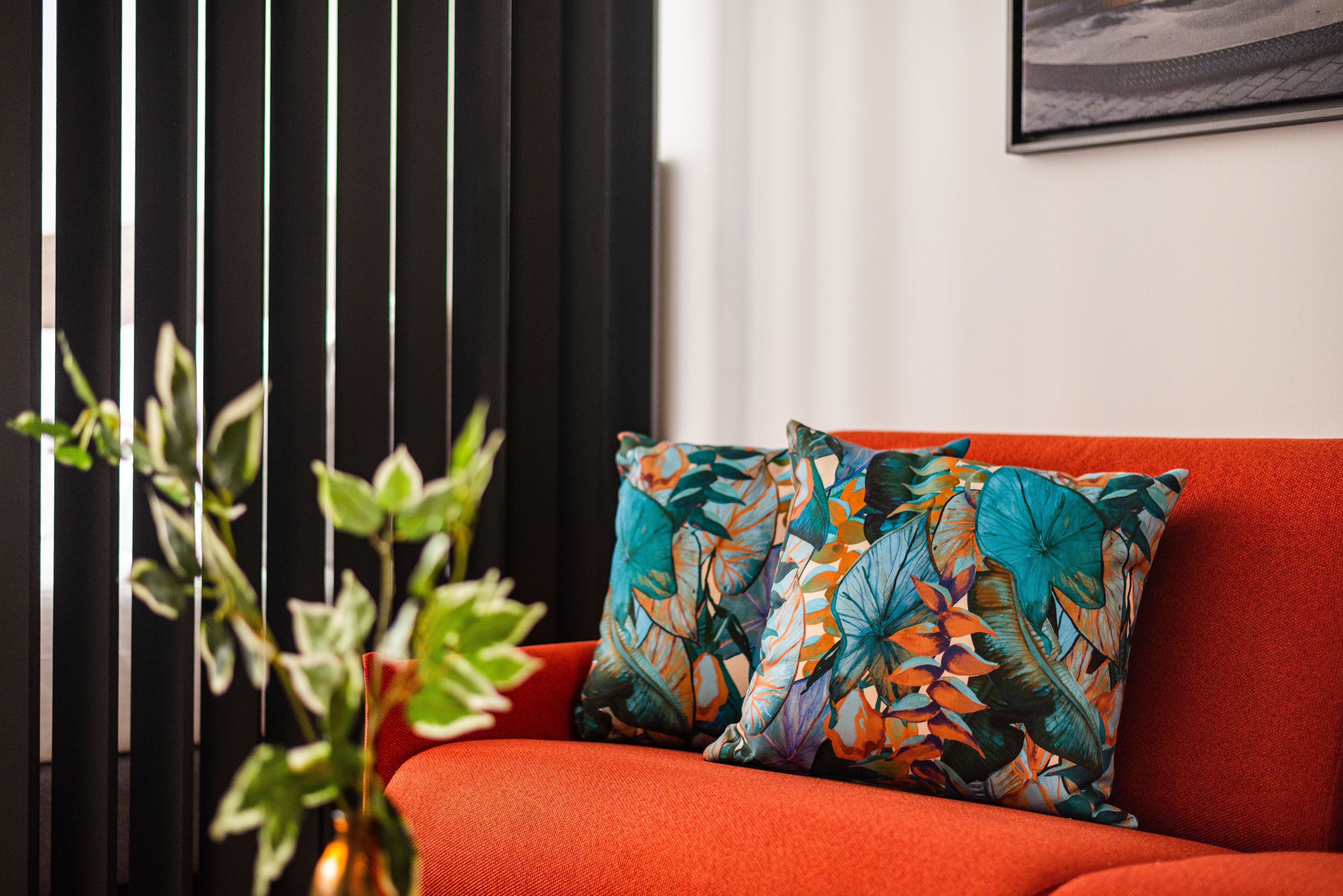 A red sofa with a colorful floral pillow, a potted plant, and a framed picture on the wall.