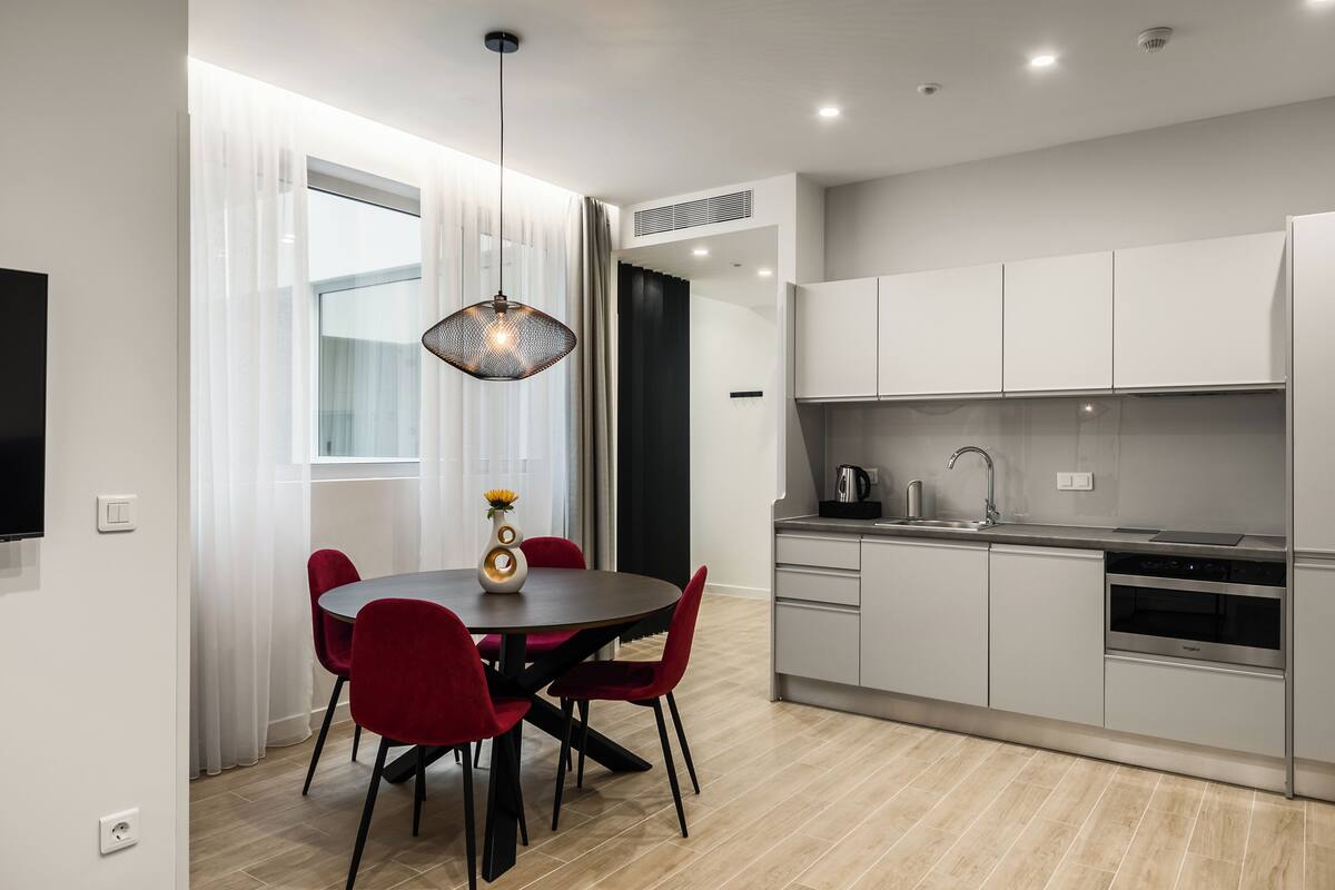 Een moderne keuken met witte kasten, een ingebouwde oven en een eethoek met een ronde tafel en rode stoelen.