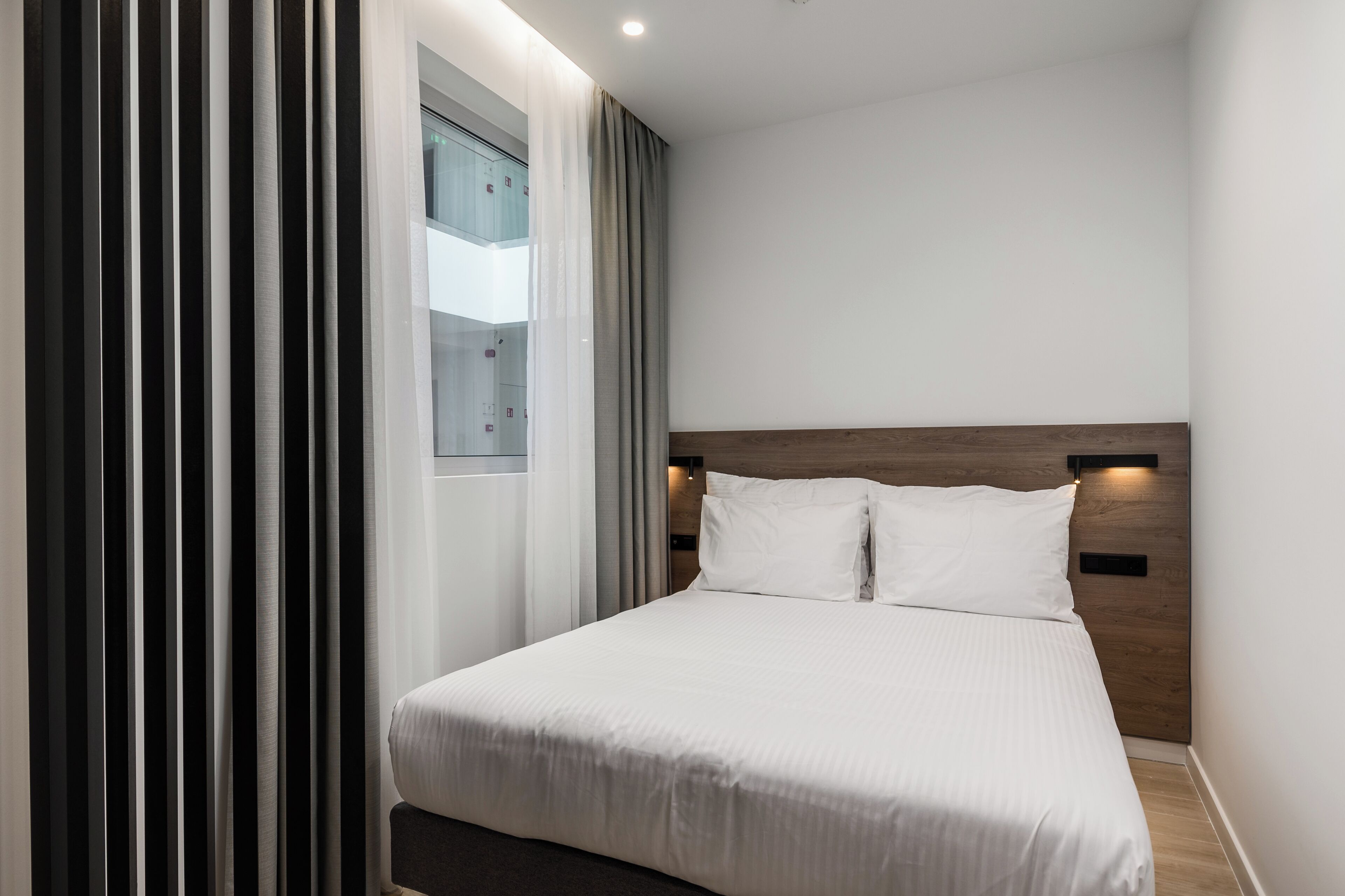 A modern hotel room with a large bed, a wooden headboard, and a view of a cityscape through a window.
