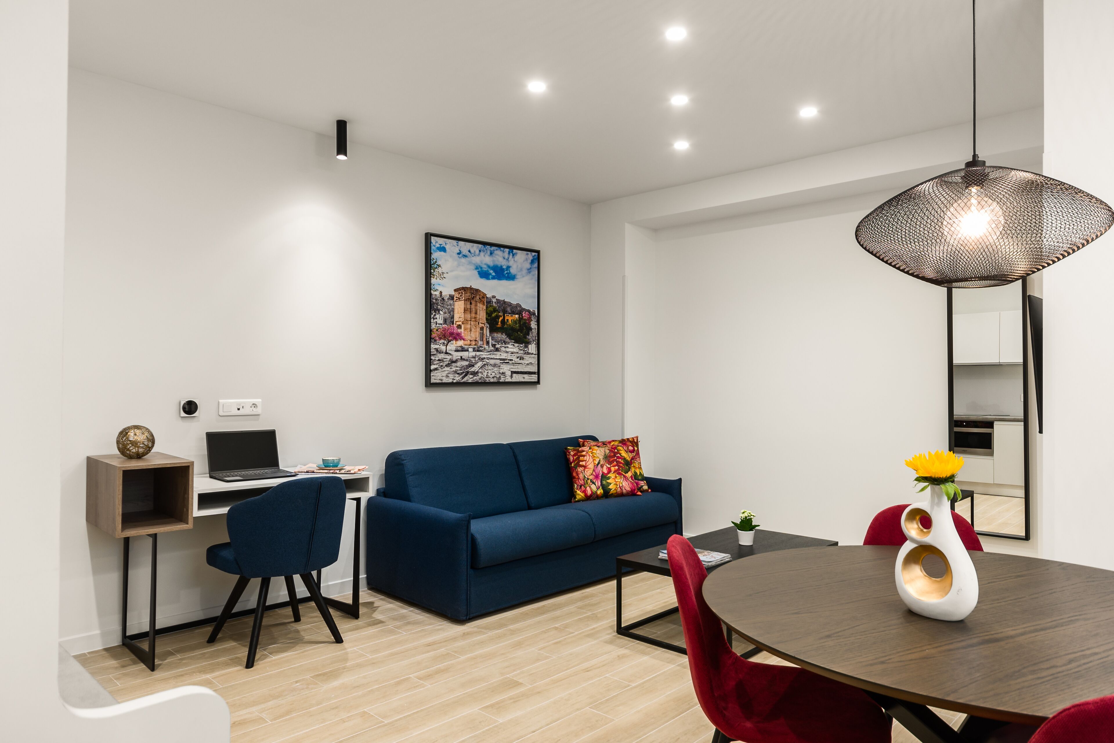 A modern living room with a blue sofa, a dining table with red chairs, a laptop on a desk, and a hanging light fixture.