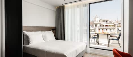 Ett modernt hotellrum med en stor säng, utsikt över staden genom en skjutdörr i glas och en matplats som syns genom dörren.