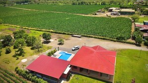 Un edificio grande con techo rojo y una piscina al frente, rodeado de campos verdes exuberantes y montañas a lo lejos.