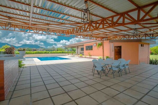 A covered outdoor area with a swimming pool, a small building, and a patio with chairs.