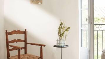 A wooden chair, a small table with a vase of plants, and a window with a view of greenery.